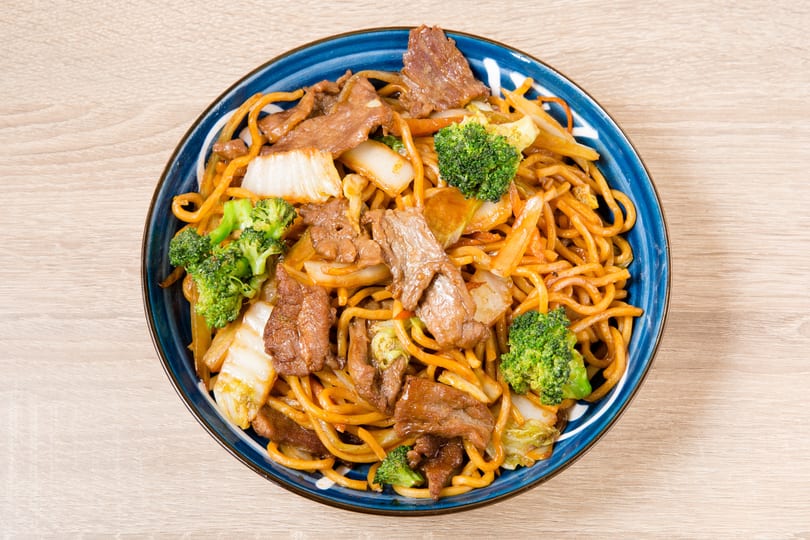 A blue bowl of beef chow mein with broccoli, cabbage, and carrots on a wooden table.