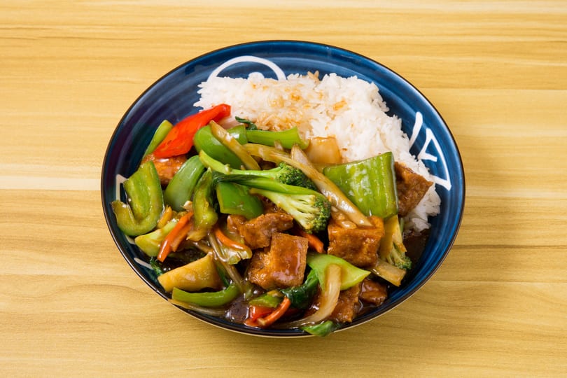 Tofu and mixed vegetables stir-fry with white rice in a blue bowl on a wooden table.