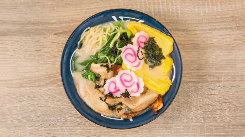 Overhead view of a ramen bowl with pork, noodles, greens, egg, and pink narutomaki.