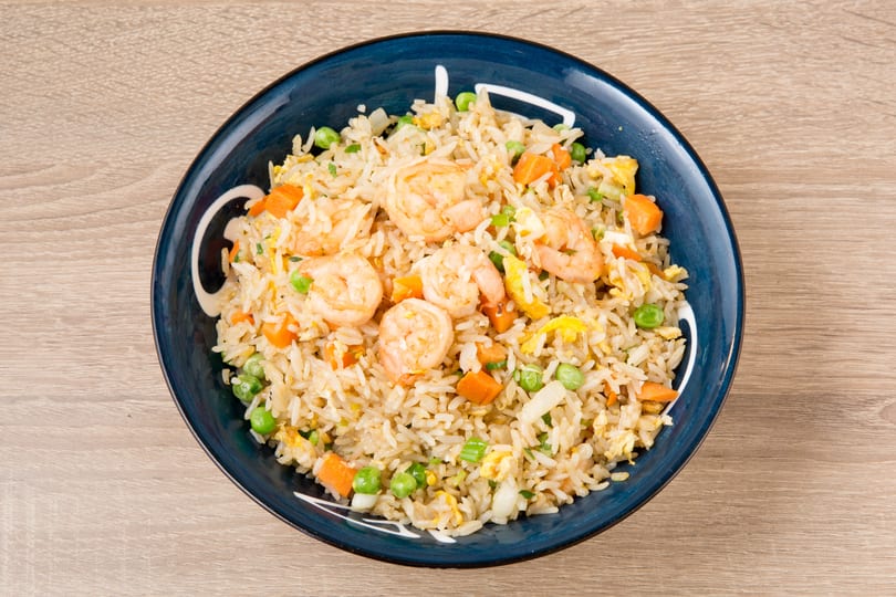 Shrimp fried rice with peas, carrots, and scrambled egg in a blue bowl on a wooden table.