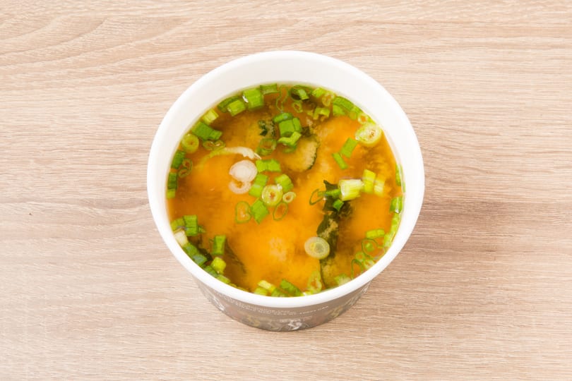 A white bowl of miso soup with green onions and seaweed sits on a light wooden table.