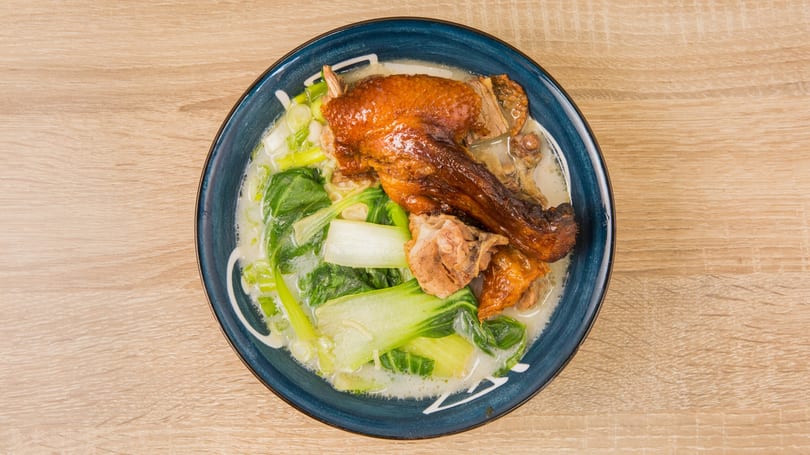 Roasted duck leg and bok choy in a creamy soup, served in a blue bowl on a wooden table.