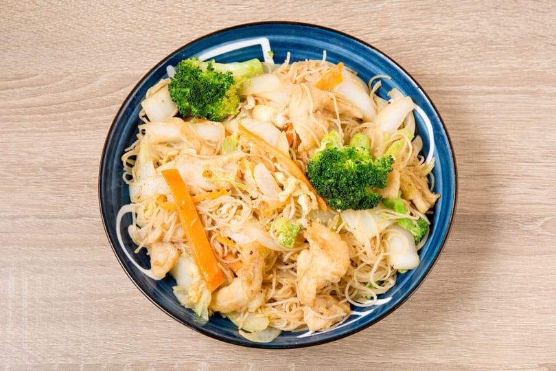 A blue bowl of Singapore noodles with chicken, broccoli, carrots, and cabbage on a wooden table.