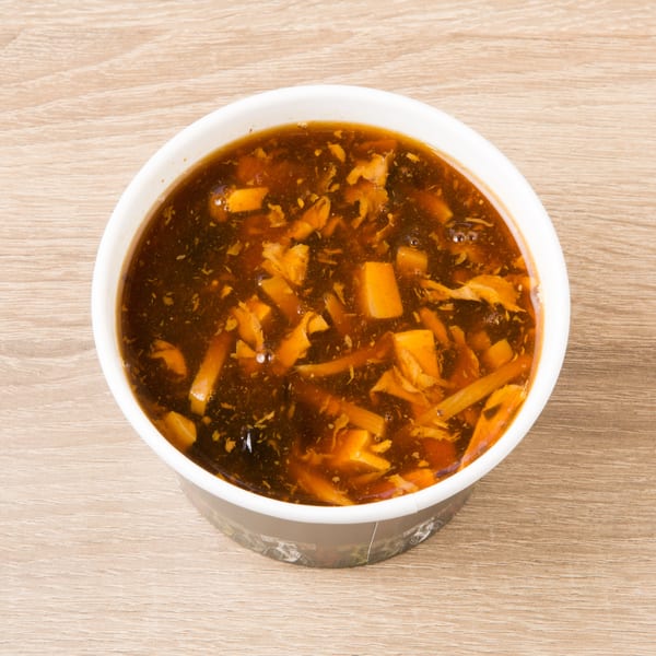 Hot and sour soup in a white and black takeout container on a light wooden table.