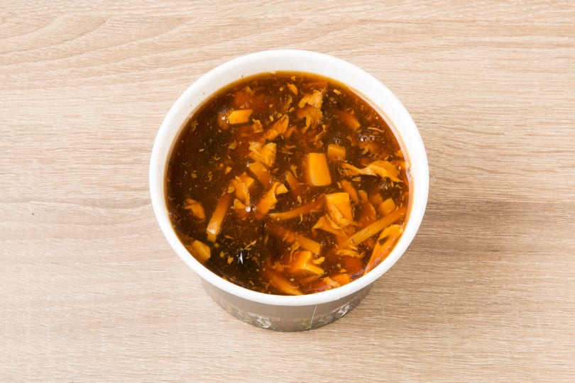 Hot and sour soup in a white and black takeout container on a light wooden table.
